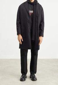 Cappotto di lana nero con rever, due bottoni e tasche laterali, indossato sopra una felpa nera e una sciarpa, abbinato a pantaloni neri e scarpe.