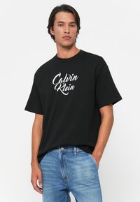 Joven con cabello oscuro que lleva una camiseta negra de Calvin Klein y jeans azul claro, de pie con una mano en el bolsillo.