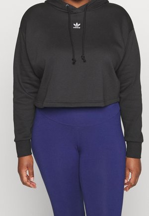 Persoon draagt een zwarte cropped Adidas hoodie met logo en koningsblauwe leggings met hoge taille, staand tegen een effen achtergrond.