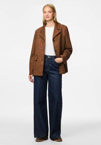 Smeđa suđena jakna s rever kragnom i zatvaranjem na dugme, nošena preko bijele bluze i tamnoplave široke traperice. Glatka tekstura i strukturirani oblik.