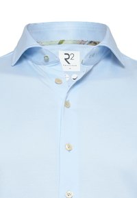 Hemelsblauw button-up shirt van een gestructureerde stof, met een bloemenbedrukte kraag, vijf knopen en een label met de tekst "R2 Amsterdam M."