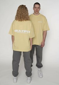 Gelbe übergroße T-Shirts mit weißem "MULTIPLY"-Logo auf der Rückseite. Kombiniert mit grauen Jogginghosen und weißen Sneakers, die einen entspannten Sitz zeigen.