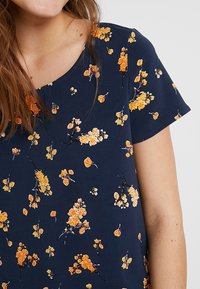 Merkkipaidassa on lyhyet hihat ja merenk Blue-kukkakuvio, joka sisältää oransseja ja valkoisia kukkia. Paita on valmistettu pehmeästä kankaasta ja siinä on pyöreä kaula-aukko.