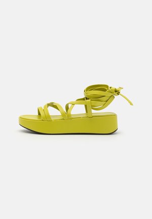 Gul platformsandal med flere tynde remme og en snor, der vikles om anklen, på en flad hvid overflade.