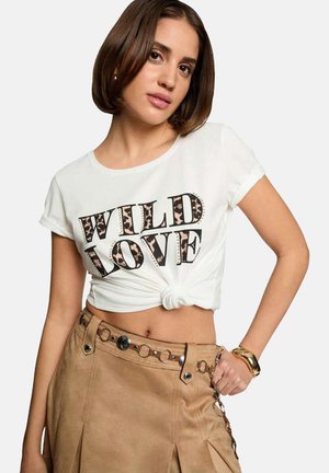 T-shirt bianco a maniche corte e corta con la scritta "WILD LOVE" in nero e dettagli in stampa leopardata. Annodato in vita, indossato con una gonna beige.