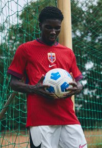 Rød fodboldtrøje med strukturerede striber, blå accenter og et nationalt emblem. Spilleren holder en hvid og blå fodbold og står ved siden af en målstang.