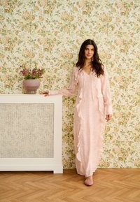 Žena v dlouhých růžových šatech s volány a sladěnými botami stojí vedle bílé krytky radiátoru s květináči, na pozadí květinová tapeta.