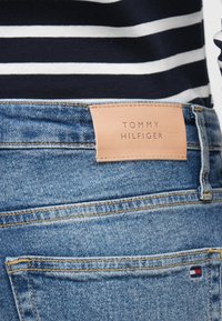 Μπλε τζιν από denim με ταμπέλα από καφέ δέρμα που γράφει "TOMMY HILFIGER." Περιλαμβάνει τυπικές πίσω τσέπες και εμφανείς λεπτομέρειες ραφής.