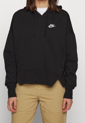 Osoba ubrana w czarną bluzę z kapturem Nike z białym logo i sznurkami, zestawioną z beżowymi spodniami, stojąca na jednolitym tle.