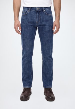 Uomo che indossa jeans blu slim fit e stivali di pelle marrone scuro, in piedi su uno sfondo bianco semplice.