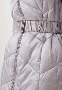 Giacca imbottita di un viola chiaro con cintura in vita, caratterizzata da motivi a diamante trapuntati, zip bidirezionale e materiale esterno lucido e liscio.