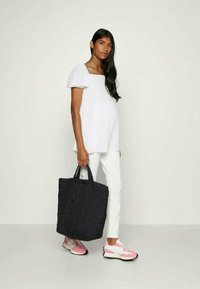 Mala tote preta com acolchoamento, segurada com uma mão, usada com uma blusa branca e calças brancas; sapatos de desporto rosa e cinza completam o visual.