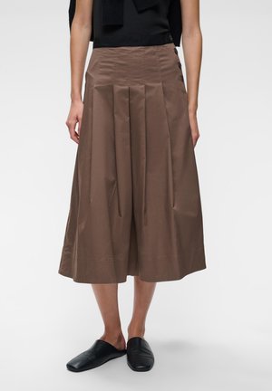 Femme portant une jupe midi plissée marron avec des détails de boutons et des chaussures noires sans lacets, debout devant un fond clair uni.