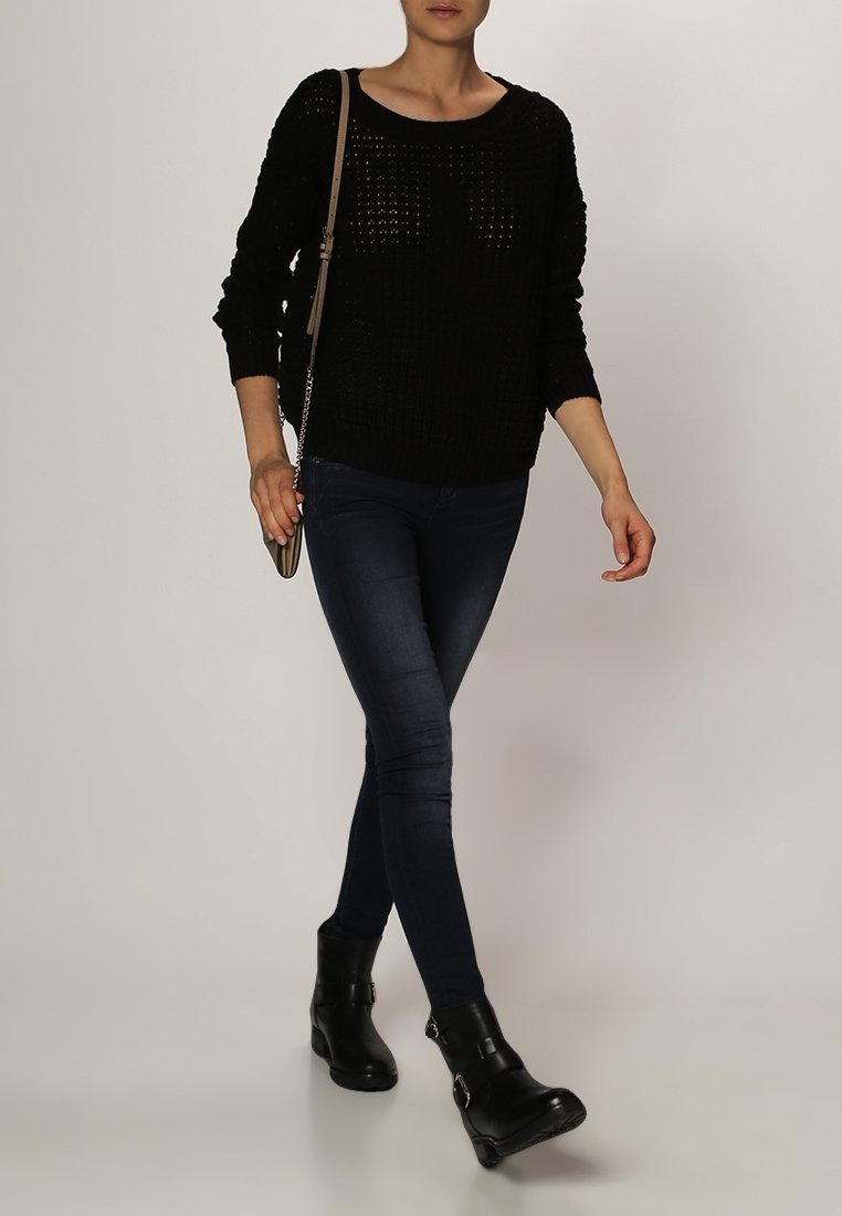 Personne marchant portant un pull noir en maille, un jean skinny foncé, des bottines noires et un sac bandoulière marron sur un fond clair.