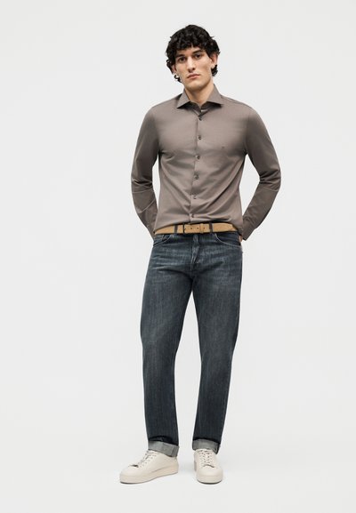 Νέος άνδρας όρθιος με τα χέρια πίσω από την πλάτη, φοράει γκρι πουκάμισο με κουμπιά, σκούρο τζιν, μπεζ ζώνη και λευκά αθλητικά παπούτσια σε απλό φόντο.