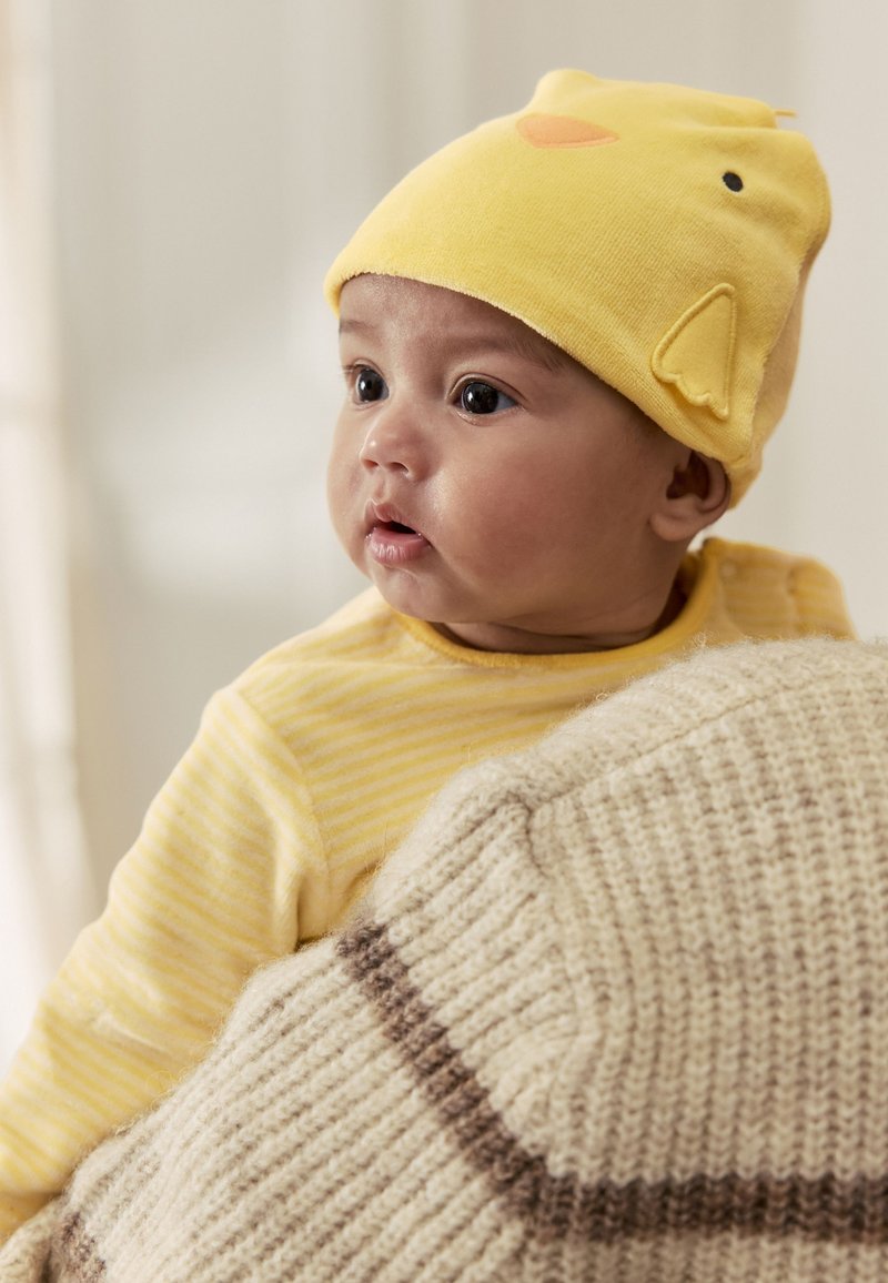 Bebé con un gorro amarillo con diseño de pollito y ropa a rayas amarillas, siendo sostenido por una persona con un suéter de punto beige.