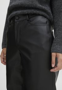 Pantaloni in ecopelle neri con una texture liscia, dotati di chiusura a bottone, tasche laterali e design a gamba dritta. Indossati con un maglione in maglia grigia.