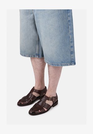 Sandalias de cuero marrón con un diseño trenzado, que presentan la punta abierta y una correa en el tobillo con hebilla. Llevadas con pantalones cortos de mezclilla de color azul claro.