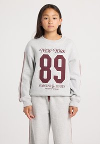 Girl wearing gray sweatshirt and sweatpants with maroon stripes, sweatshirt text reads “New York 89 Forever & Always The City of Dreams.”