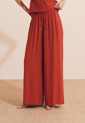 Personne portant un pantalon ample, large et rouge rouille avec une taille élastique et un cordon, debout pieds nus sur un sol texturé.