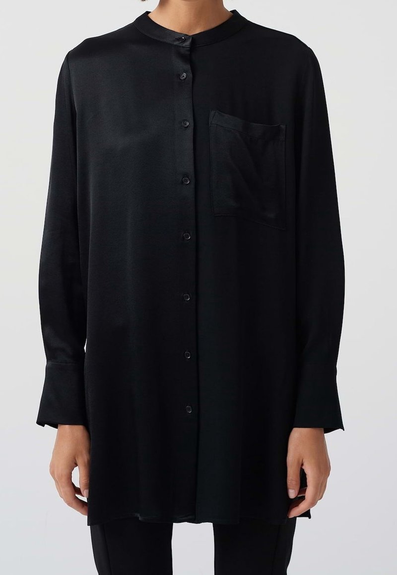Femme portant une chemise noire à manches longues boutonnée, avec un col rond et une poche poitrine, debout les bras détendus le long du corps.
