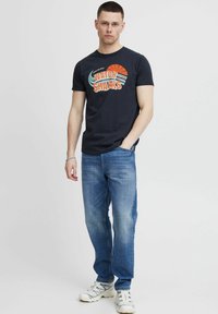 Navy T-Shirt mit einem bunten Grafikdesign und dem Text "WATCH SHARKS", kombiniert mit blauen Jeans und weißen Sportschuhen.