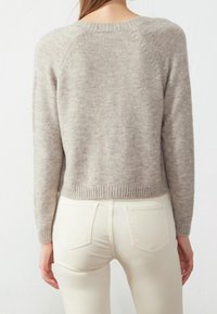 Hellgrauer Strickpullover mit langen Ärmeln und geripptem Saum; über weißen Slim-Fit-Hosen getragen. Hat einen runden Halsausschnitt und Raglan-Nähte.