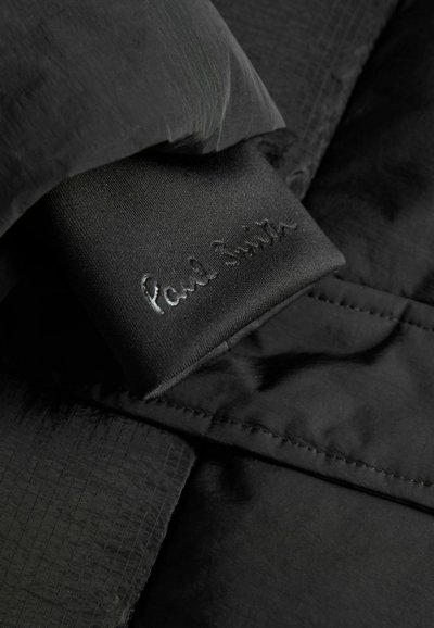 Tuņa tuvplānā ir melna auduma apģērba etiķete ar iepresētu "Paul Smith" tekstu, piestiprināta pie teksturēta un stitched melna apģērba.