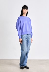 Sudadera morada de gran tamaño con hombros caídos, combinada con jeans rectos de color azul claro y mocasines negros, sobre un fondo neutro.