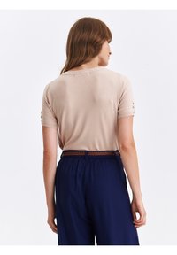 Beżowy sweter z krótkim rękawem i ściągaczem przy dekolcie, z guzicznymi akcentami na rękawach, w połączeniu z granatowymi spodniami i plecionym paskiem.