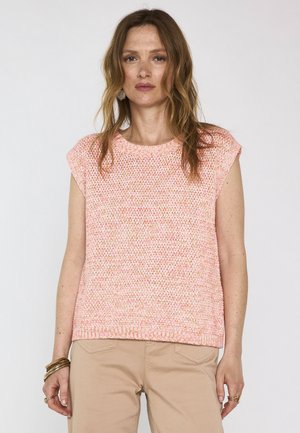 Donna con capelli castano chiaro ondulati che indossa un top rosa lavorato a maglia senza maniche e pantaloni beige, in piedi davanti a uno sfondo chiaro uniforme.