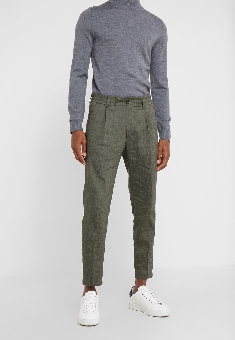 Personne portant un pantalon fuselé vert olive, un pull gris à col roulé manches longues et des baskets blanches, debout devant un fond uni.