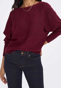 Burgundský pletený svetr s volným střihem, širokým lodičkovým výstřihem a vroubkovanými manžetami, doplněný tmavými džíny s klasickým pětivreckovým designem.