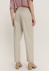 Una donna in pantaloni tapered beige a vita alta con cintura elastica e scarpe con tacco a blocco beige, in piedi contro uno sfondo neutro.