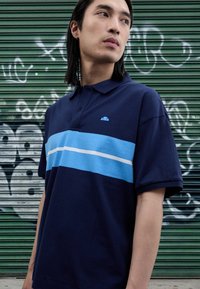 Marinblå pikétröja med ett horisontellt mönster i blått och ljusblått. Kortärmad, med krage och en liten logotyp på bröstet.