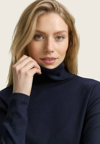 Femme blonde aux yeux bleus portant un col roulé bleu marine, tenant le col de sa main gauche contre un fond clair.