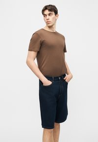 Νεαρός άνδρας ντυμένος με καφέ κοντομάνικο πουκάμισο και σκούρες μπλε τζιν βερμούδες, στέκεται με τα χέρια στις τσέπες, κοιτάζοντας προς τα αριστερά σε απλό φόντο.