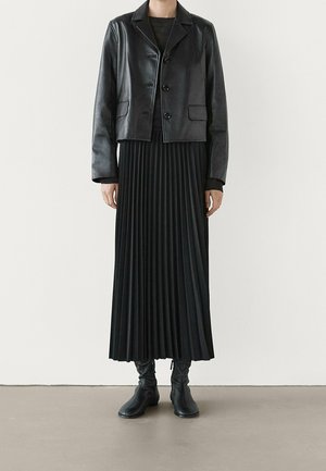 Personne portant une veste en cuir noire raccourcie, une longue jupe plissée noire et des bottines noires, debout contre un mur blanc.