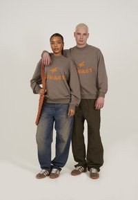 Hanorac maro cu text portocaliu "LEGACY" și o stea. Modelul poartă blugi largi albaștri, iar celălalt poartă pantaloni de culoare verde oliv, amândoi având încălțăminte cu dungi.