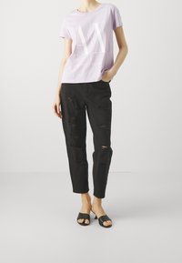 Camiseta de algodón lavanda con un gráfico blanco del logo "AX", combinada con jeans de mezclilla rasgados negros y sandalias de slide negras.