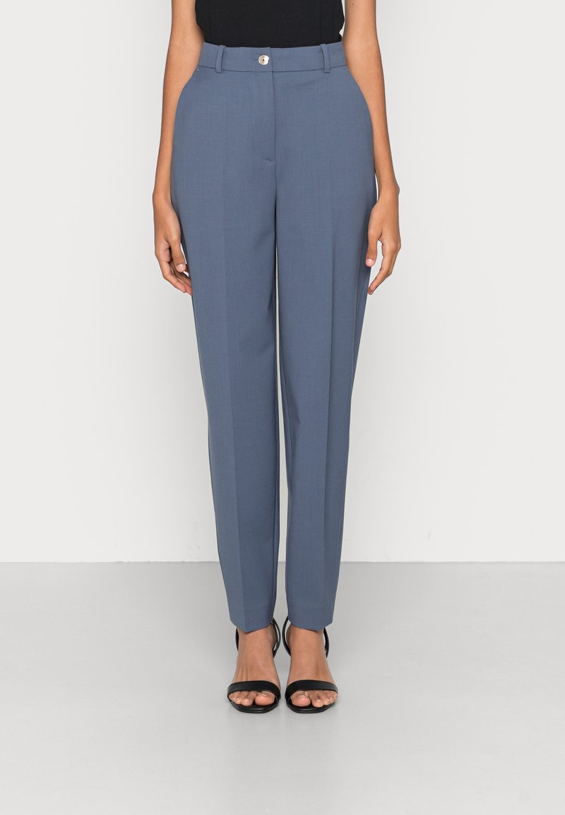 Kobieta w niebieskich, dopasowanych spodniach z przodu zapinanych na guzik oraz czarnych, odkrytych szpilkach, stojąca na jednolitym białym tle.