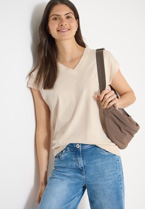 Donna con lunghi capelli scuri che indossa una maglietta beige a maniche corte con scollo a V e jeans blu, che tiene la tracolla di una borsa marrone su sfondo bianco.