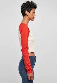 Rotes und cremefarbenes Crop-Top mit langen Ärmeln, aus weichem Stoff hergestellt. Hat einen runden Ausschnitt. Kombiniert mit blauen Jeans.