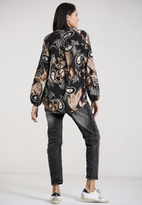 Schwarzbluse mit floralen Mustern und Ballonärmeln, kombiniert mit grauen Jeans und weißen Sneakers. Die Bluse hat große Paisleymuster in Beige und Braun.