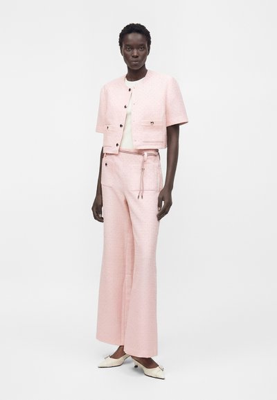 Femme debout portant une veste courte rose pâle à manches courtes et un pantalon large assorti, avec des chaussures blanches à talons bas.