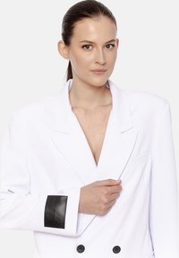 Kobieta z ciemnymi, upiętymi włosami, nosząca obszerne, białe, podwójnie zapięte żakiet z czarną łatką na mankiecie, trzymająca klapę pod światło na jasnym tle.