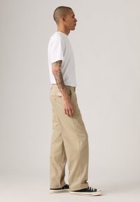 Man with tattoos wearing a white t-shirt, beige wide-leg pants, and black sneakers standing in profile against a plain background.