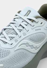 Baskets de course gris clair avec un upper en mesh texturé, des lacets blancs et une semelle verte discrète. Logo de marque bien visible sur le côté.