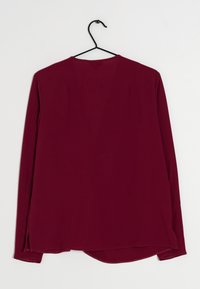 Blusa bordeaux a maniche lunghe con scollatura a V, realizzata in tessuto leggero, caratterizzata da fessure laterali e un orlo leggermente curvato. Appesa a un gancio nero.