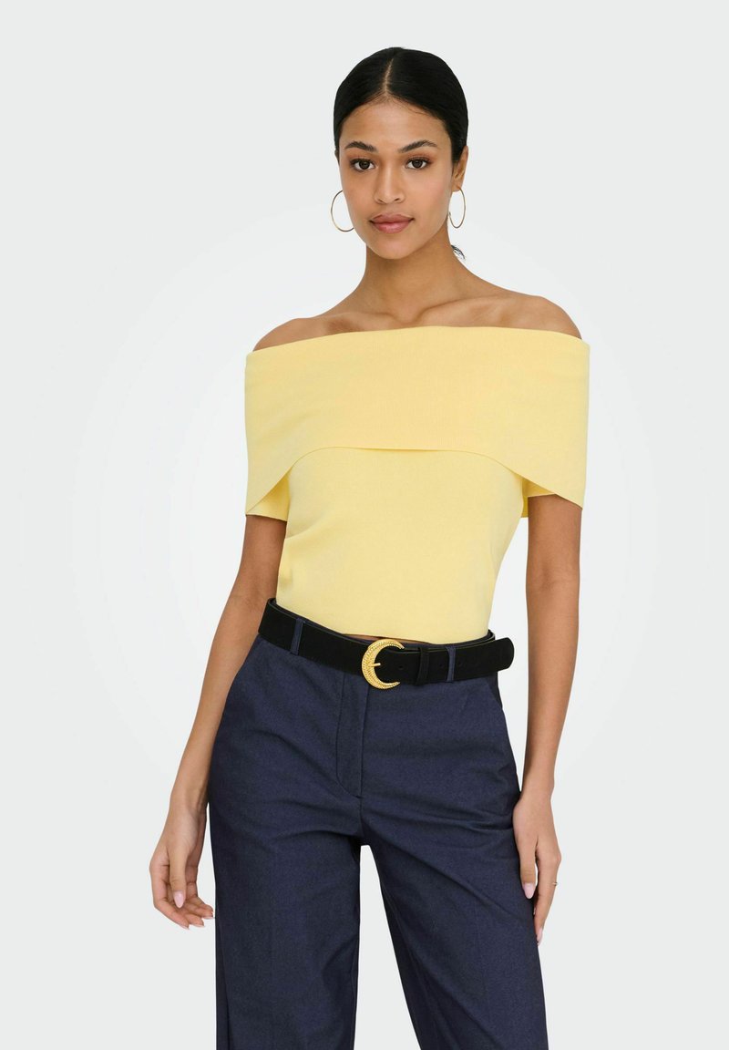 Femme portant un haut jaune à épaules dénudées, un pantalon taille haute bleu marine avec une ceinture noire ornée d'une boucle dorée, et des boucles d'oreilles créoles dorées.